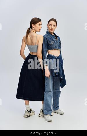 Due bellissime sorelle si mettono in posa uno contro l'altro, mettendo in risalto i loro esclusivi outfit in denim in uno studio. Foto Stock