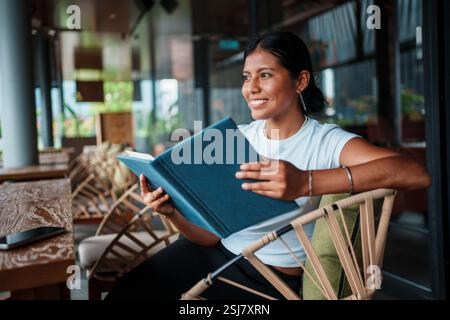 Una donna sorridente ama leggere un libro mentre si rilassa in un caffè accogliente e confortevole Foto Stock