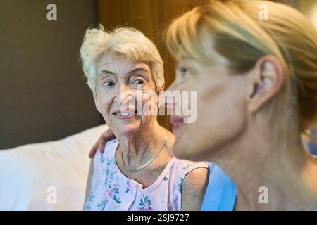 Una donna anziana vestita con un top floreale sorride, seduta in una stanza illuminata, mentre una giovane infermiera offre sostegno e compagnia, Foto Stock