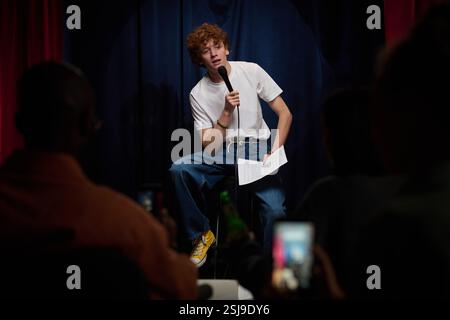 Persona che esegue una commedia in piedi sul palco tenendo in mano il microfono e la sceneggiatura. I partecipanti del pubblico sono visibili con i telefoni che catturano l'attimo Foto Stock