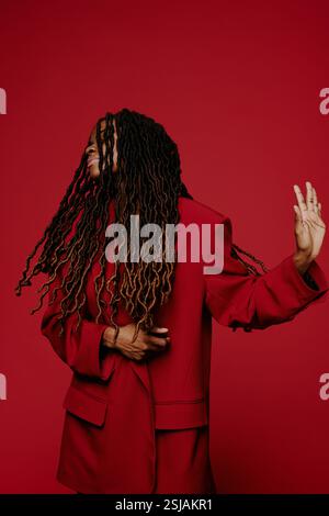 Donna nera con lunghi dreadlock che ballano e gestiscono su sfondo rosso intenso e semplice, indossando un elegante abito rosso Foto Stock