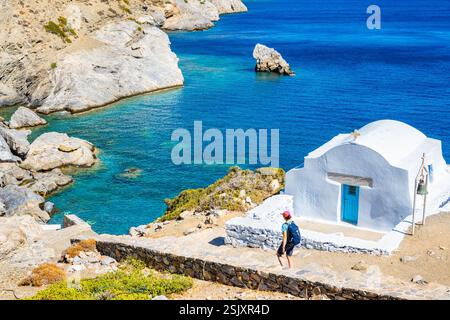 Donna turistica con zaino che cammina sul sentiero per la spiaggia di Agia Anna e passa accanto alla piccola cappella greca bianca, all'isola di Amorgos, alle Cicladi, in Grecia Foto Stock