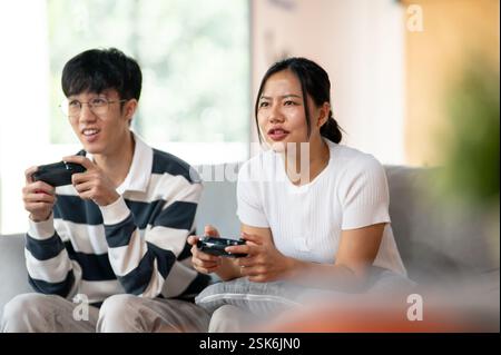 Due amici asiatici, un maschio e una femmina, giocano con entusiasmo a un videogioco con joystick sul divano in salotto, per divertirsi insieme a casa. Foto Stock
