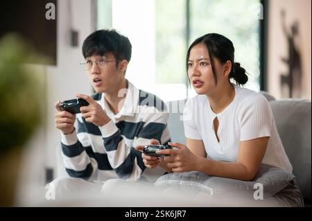 Due amici asiatici, un maschio e una femmina, giocano con entusiasmo a un videogioco con joystick sul divano in salotto, per divertirsi insieme a casa. Foto Stock