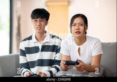 Due amici asiatici, un maschio e una femmina, giocano con entusiasmo a un videogioco con joystick sul divano in salotto, per divertirsi insieme a casa. Foto Stock