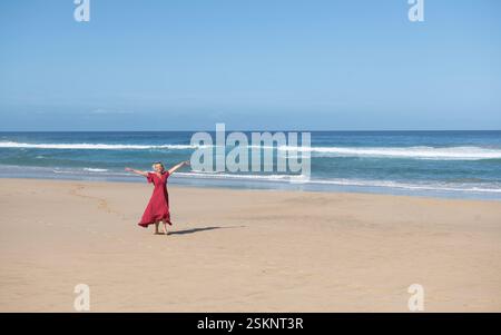 Donna felice con un vestito rosso in piedi sulla spiaggia con le braccia allungate, spiaggia la Cofete, Fuerteventura, Isole Canarie, Spagna Foto Stock