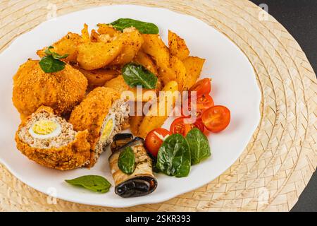Una deliziosa sistemazione comprende croccanti pepite di pollo, patate arrostite e pomodori ciliegini freschi con spinaci, serviti elegantemente su un piatto bianco Foto Stock