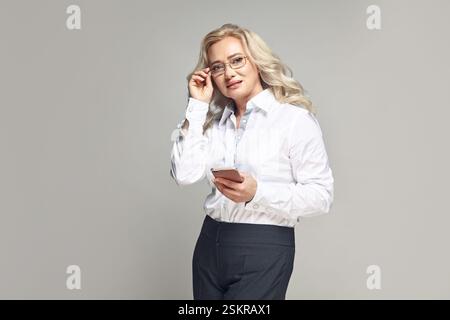 Donna matura in affari con occhiali che tiene il telefono in mano su grigio isolato. Foto Stock