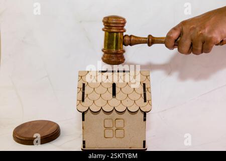 Una mano regge un martelletto di legno sopra un piccolo modello di casa in legno, simboleggiando un processo di asta immobiliare. Foto Stock