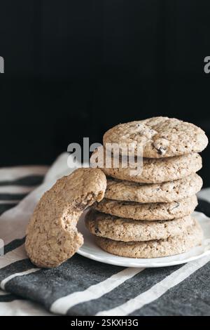 Un piatto di biscotti senza glutine con scaglie di cioccolato impilati ordinatamente, con un biscotto che mostra un morso estratto, e posti su un panno a strisce contro un dorso scuro Foto Stock