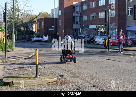 Punto di vista posteriore di un uomo anziano che guida uno scooter mobile su una strada principale in una zona periferica di Shepperton Surrey Inghilterra Regno Unito Foto Stock