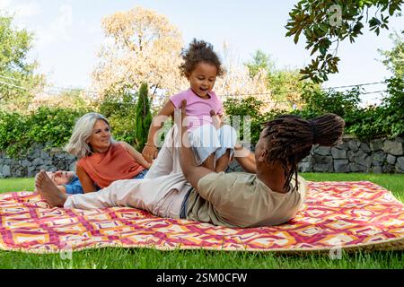 Un padre amorevole solleva sua figlia in aria mentre gioca su una coperta da picnic all'aperto. I nonni si rilassano sullo sfondo, godendosi un caldo e jo Foto Stock