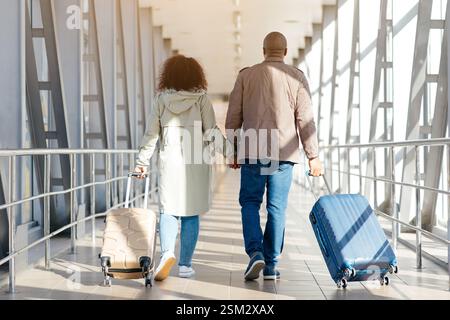 Romantica coppia africana che cammina con le valigie nel terminal dell'aeroporto, si tiene per mano e sorride, vista posteriore con spazio libero Foto Stock