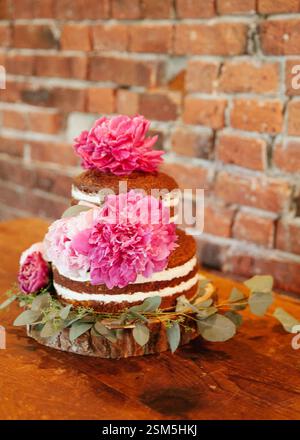 Una torta a tre livelli splendidamente lavorata presenta peonie rosa e vegetazione fresca su una superficie rustica in legno. Foto Stock
