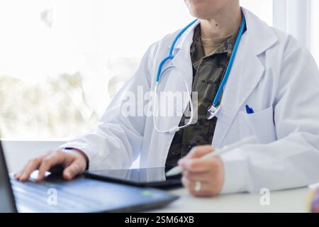 Un medico militare che indossa un cappotto bianco sopra un'uniforme mimetica funziona su un computer portatile e un tablet in un luminoso ufficio medico. Il professionista ha una stint Foto Stock