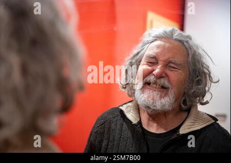 Un uomo anziano con capelli ricci grigi e barba ride gioiosamente mentre guarda il suo riflesso nello specchio, davanti a una parete rossa vibrante. Foto Stock