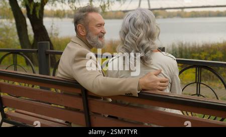 Back view felice anziano maturo 60s marito moglie abbracciare sulla panchina all'aperto sano pensione coppia donna mezza età uomo parlare ridere rilassarsi godere romantico Foto Stock