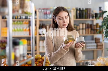 Giovane donna che tiene in mano due lattine di conserve di latta, sceglie e fa shopping intorno a pate per dei panini Foto Stock