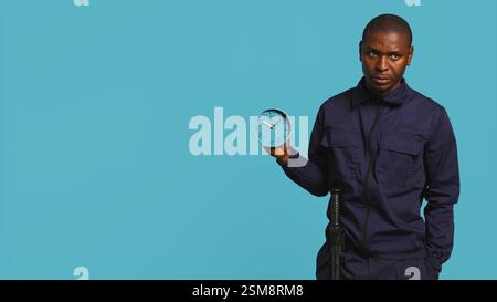 Guardia di sicurezza che tiene l'orologio, fa uscire la gente dalla proprietà, isolata sullo sfondo dello studio. Uomo in uniforme che mostra ai visitatori il tempo passa, si prepara a chiudere la postazione, telecamera B. Foto Stock
