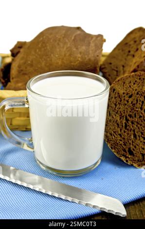 Latte in un calice di vetro con pane di segale in un cestello di vimini e un coltello su un tovagliolo blu e un bordo di legno isolato su sfondo bianco Foto Stock