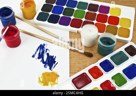 Due scatole di vernice, tre spazzole, vasi con gouache, un foglio di carta con un'immagine su una tavola di legno Foto Stock