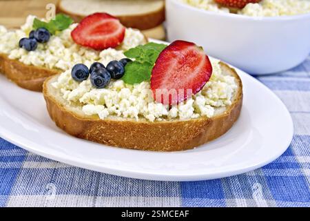Fette di pane con crema di cagliata, menta, mirtilli e fragole, una ciotola di formaggio fresco, pane su tavola su uno sfondo con scacchi blu Foto Stock