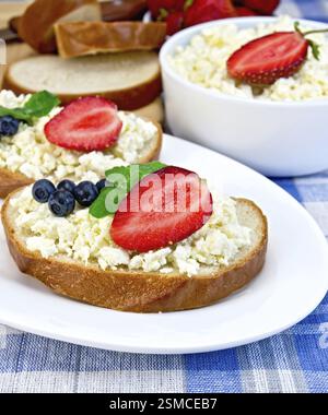 Fette di pane con crema di cagliata, mirtilli e fragole, menta, una ciotola di formaggio fresco, pane su un tavolo su uno sfondo di scacchi blu Foto Stock
