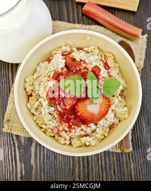 Farina d'avena in un recipiente di bambù con salsa di fragole e rabarbaro su un tovagliolo di burlap, cucchiaio, latte in una caraffa di vetro, rabarbaro contro le scure tavole sopra Foto Stock