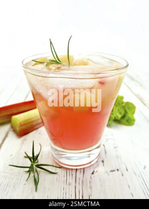 La limonata con il rabarbaro e il rosmarino in un vetro, i gambi e le foglie di rabarbaro sullo sfondo la luce di tavole di legno Foto Stock