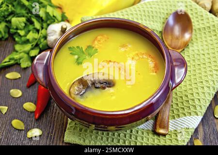Zuppa-purea di zucca con gamberi e funghi in un recipiente di argilla con un cucchiaio sull'asciugamano, zucca, prezzemolo, zenzero, cialde di peperoncino, semi di zucca su t Foto Stock