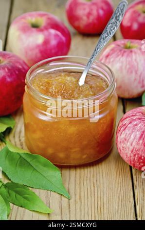 La marmellata di mele in un vasetto di vetro con un cucchiaio, fresche Mele rosse, ramoscelli con foglie su un pannello di legno Foto Stock