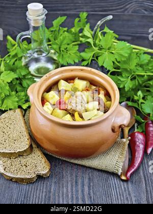 Arrosto con pollo, patate, zucca e peperoni in un vaso di argilla su un tovagliolo di tela ruvida, pane e prezzemolo al buio su un pannello di legno Foto Stock