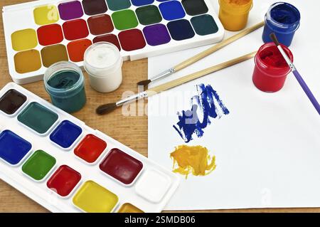 Due scatole di vernice, tre spazzole, vasi con gouache, un foglio di carta con un'immagine su una tavola di legno Foto Stock