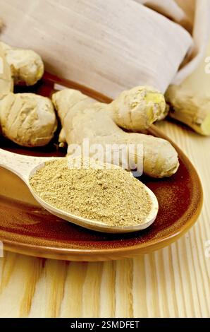 Radici di zenzero fresco, un cucchiaio di legno con una polvere di zenzero su ceramica, stoffa contro un pannello di legno Foto Stock