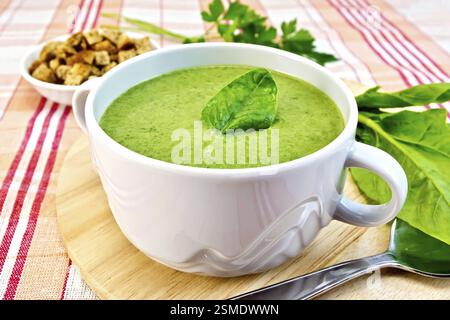 Zuppa verde purea in una ciotola con il verde di spinaci in foglia, cucchiaio sul bordo, crostini, prezzemolo su uno sfondo di stoffa Foto Stock