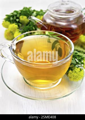 Il tè alle erbe in un bicchiere di vetro, teiera, fiori freschi, Rhodiola rosea sulla luce di sfondo di tavole di legno Foto Stock