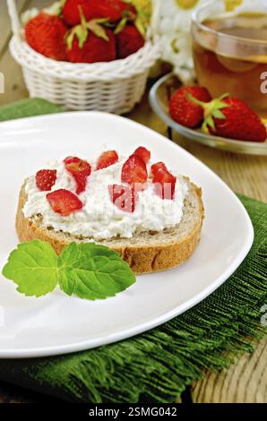 Fetta di pane francese con crema di ricotta, fragola e menta su una piastra e un tovagliolo, cesto con frutti di bosco, coppa di vetro con tè su una tavola di legno Foto Stock