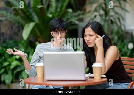 Due asiatici stressati e frustrati sono seduti di fronte a un notebook, lottando con il loro progetto, affrontando un problema durante una riunione informale Foto Stock