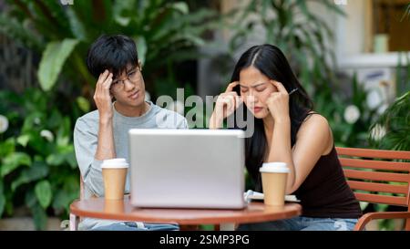 Due asiatici stressati e frustrati sono seduti di fronte a un notebook, lottando con il loro progetto, affrontando un problema durante una riunione informale Foto Stock