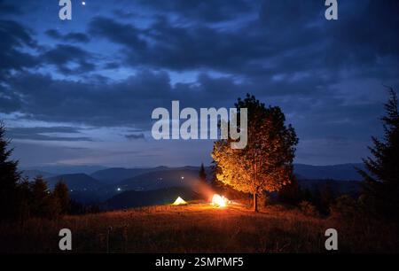 Due tende illuminate e il fuoco creano un caldo bagliore accanto all'albero sotto il cielo azzurro con nuvole sparse. L'orizzonte montuoso sullo sfondo aggiunge profondità alla tranquilla scena notturna del campeggio. Foto Stock