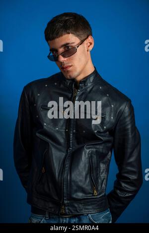 Giovane uomo alla moda con occhiali da sole e giacca in pelle nera in posa su un vivace sfondo blu Foto Stock