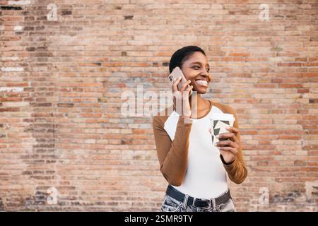 Donna afroamericana sorridente che fa una telefonata e tiene in mano una tazza di caffè usa e getta mentre si trova contro un muro di mattoni Foto Stock