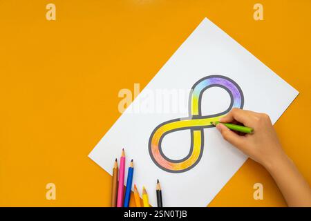 Una mano che incolla il simbolo dell'infinito arcobaleno su un foglio di carta bianco con matite colorate sparse intorno. Concetto di consapevolezza dell'autismo. Foto Stock