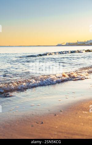 Le onde si infrangono dolcemente sulla spiaggia sabbiosa al tramonto vicino a una città costiera in una serata tranquilla Foto Stock
