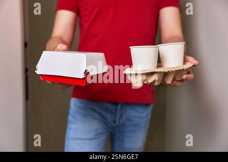 addetto alle consegne in uniforme rossa che consegna al cliente una scatola per alimenti e tazze di carta davanti alla porta d'ingresso Foto Stock