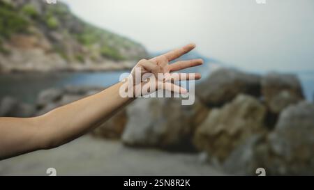 Mano dell'uomo con quattro dita sollevate dal mare, mostrando un braccio su una spiaggia panoramica all'aperto con costa rocciosa e acqua blu. Foto Stock