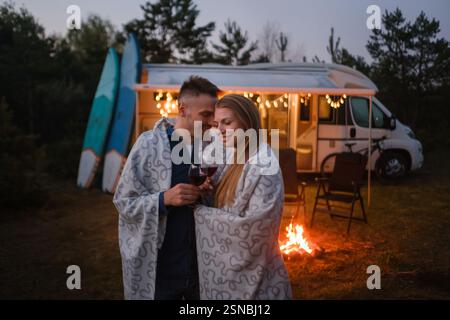 Una coppia gode di una calda serata avvolta da coperte, tenendo in mano bicchieri di vino davanti a un fuoco da campeggio e un camper, circondato da alberi e soffice mosso Foto Stock