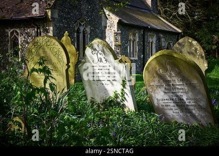 Lapidi sepolcrali storiche erose in un cimitero di campagna inglese con una chiesa parrocchiale in pietra di flint sullo sfondo Foto Stock