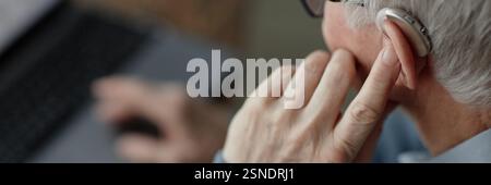 Immagine dell'intestazione del sito Web di anziani mans messa a punto dell'apparecchio acustico dietro l'orecchio premendo il dito sul dispositivo per controllare il volume del suono in ambienti chiusi, uso della tecnologia assistiva, spazio di copia Foto Stock