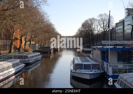 Canale di Amsterdam con tour in barca e case galleggianti in una giornata di sole. Gli alberi fiancheggiano l'acqua, riflettendo il fascino dei corsi d'acqua della città. Foto Stock